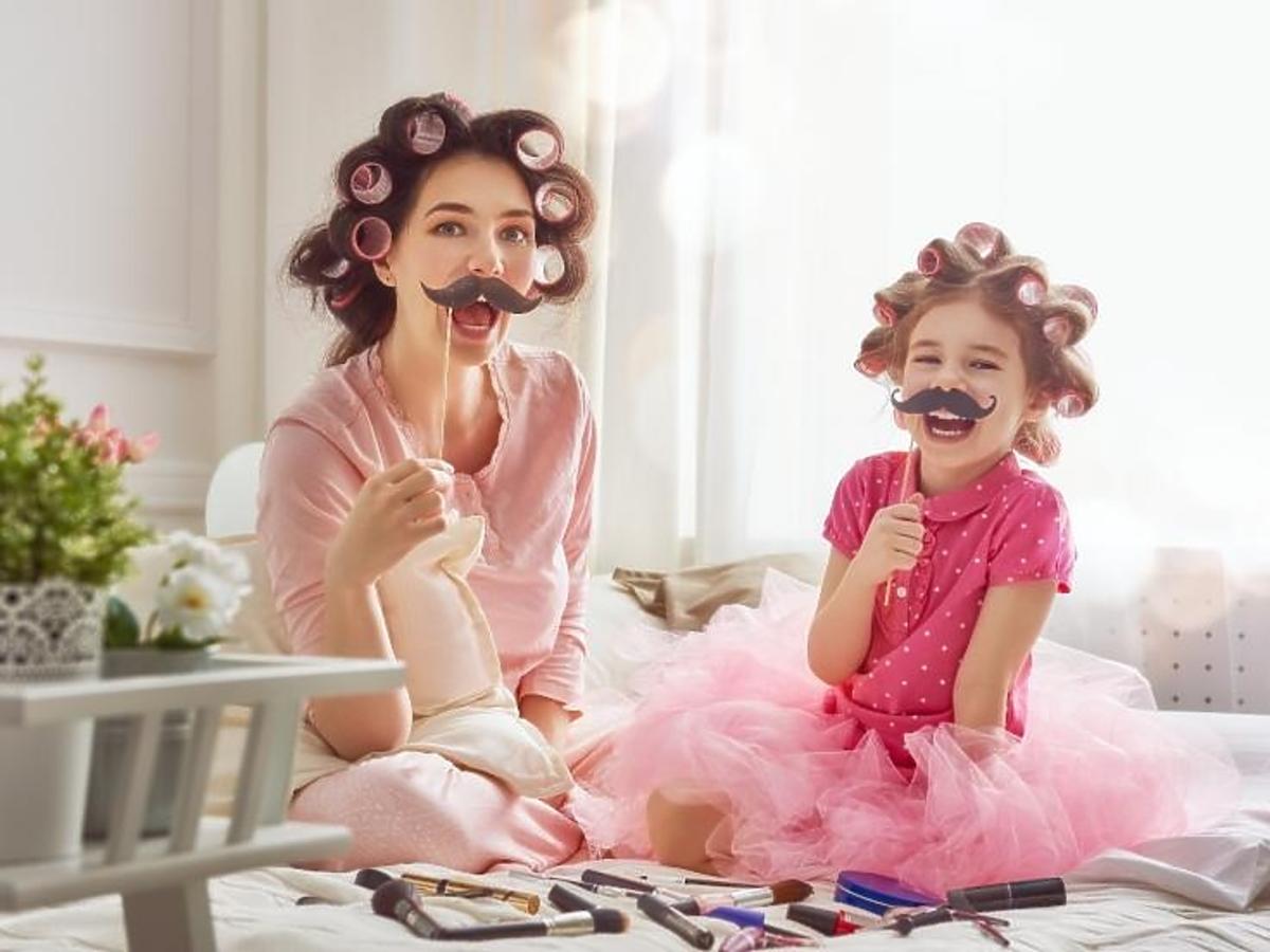 Mama siedzi na łóżku z córką i bawią się sztucznymi wąsami