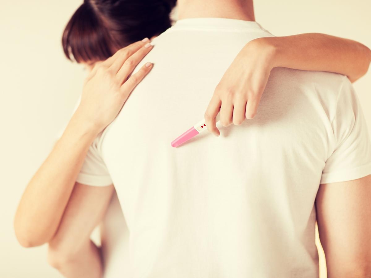 Kobieta przytula mężczyznę trzmając w ręku test ciążowy.
